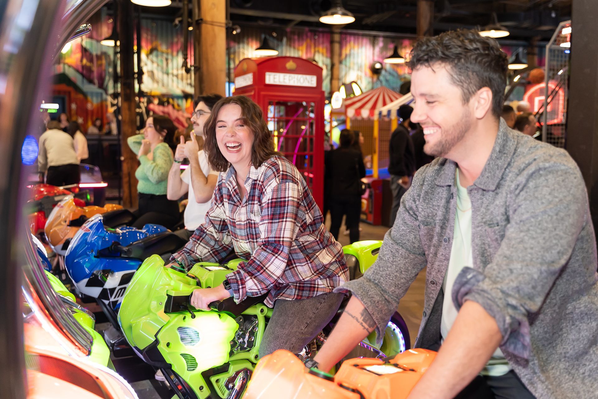 Couple laughing while playing a motorcycle arcade game in a brightly lit arcade.