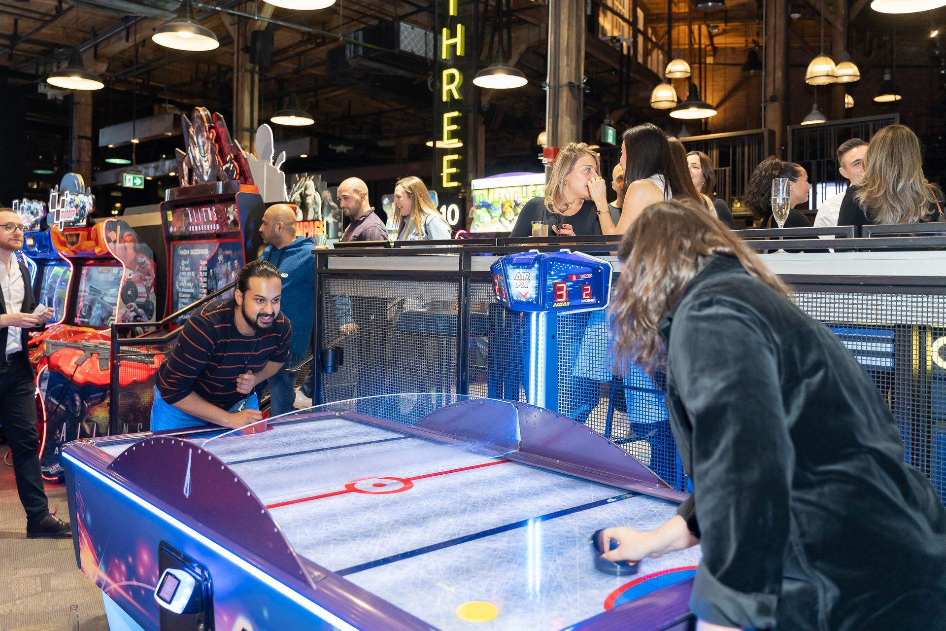 People playing air hockey in an arcade with other people and games in the background.