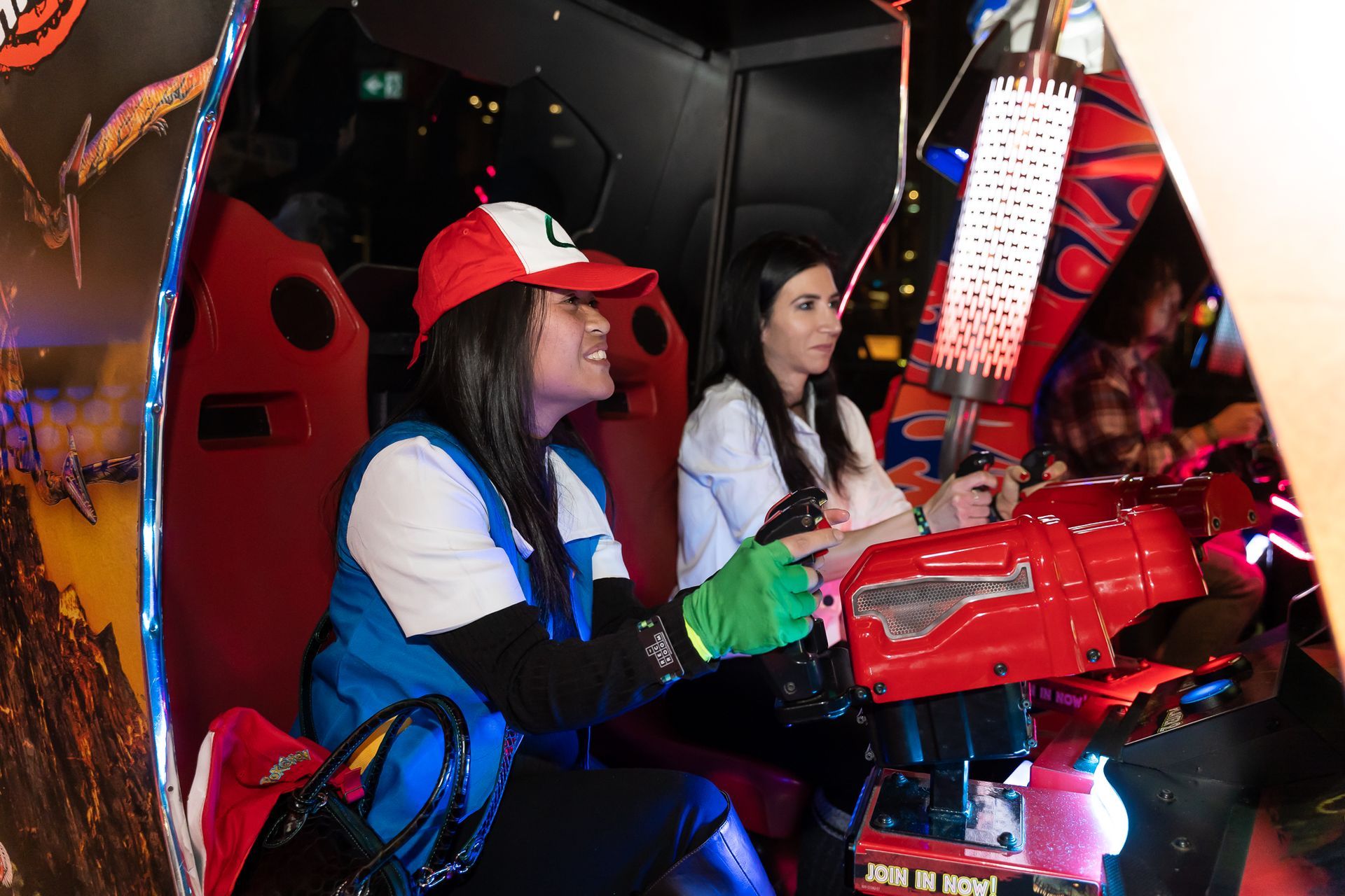 Two people playing a red arcade game. One wears an Ash Ketchum costume, smiling. Inside a dark arcade.