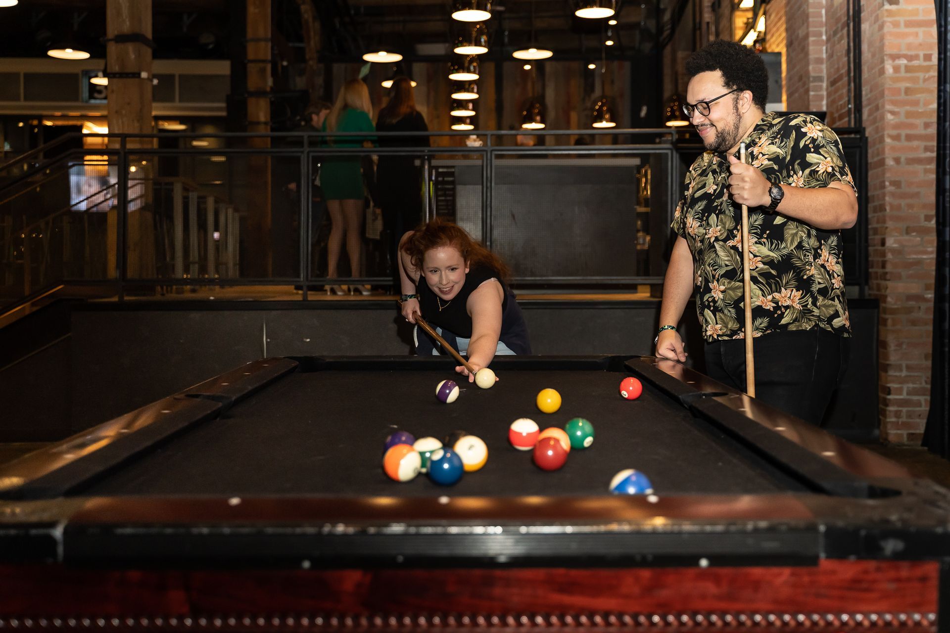 Two people playing pool in a bar. One prepares to take a shot, the other watches, smiling.