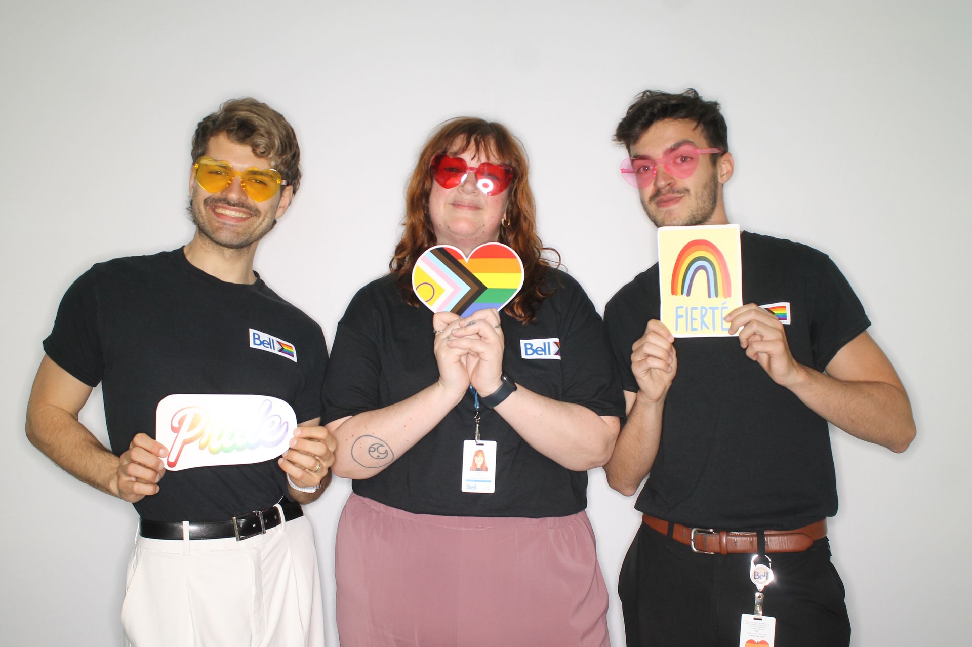 Three people wearing colored sunglasses hold Pride-themed signs, smiling. Plain white background.