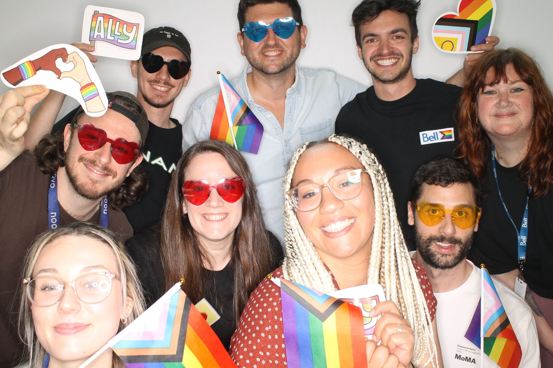Group of people smiling, holding pride flags and props, some wearing heart-shaped sunglasses. White backdrop.
