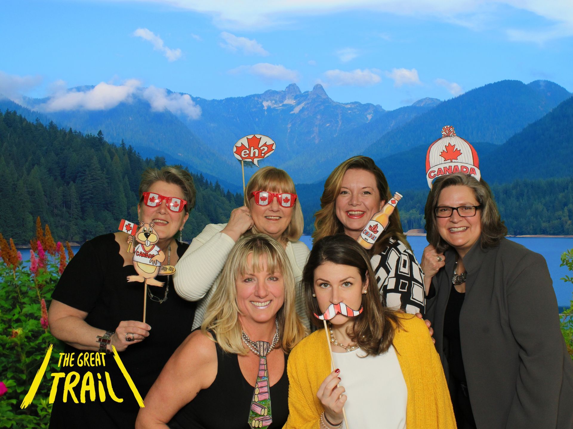 Group of women posing with Canadian-themed props in front of a mountain and lake background.