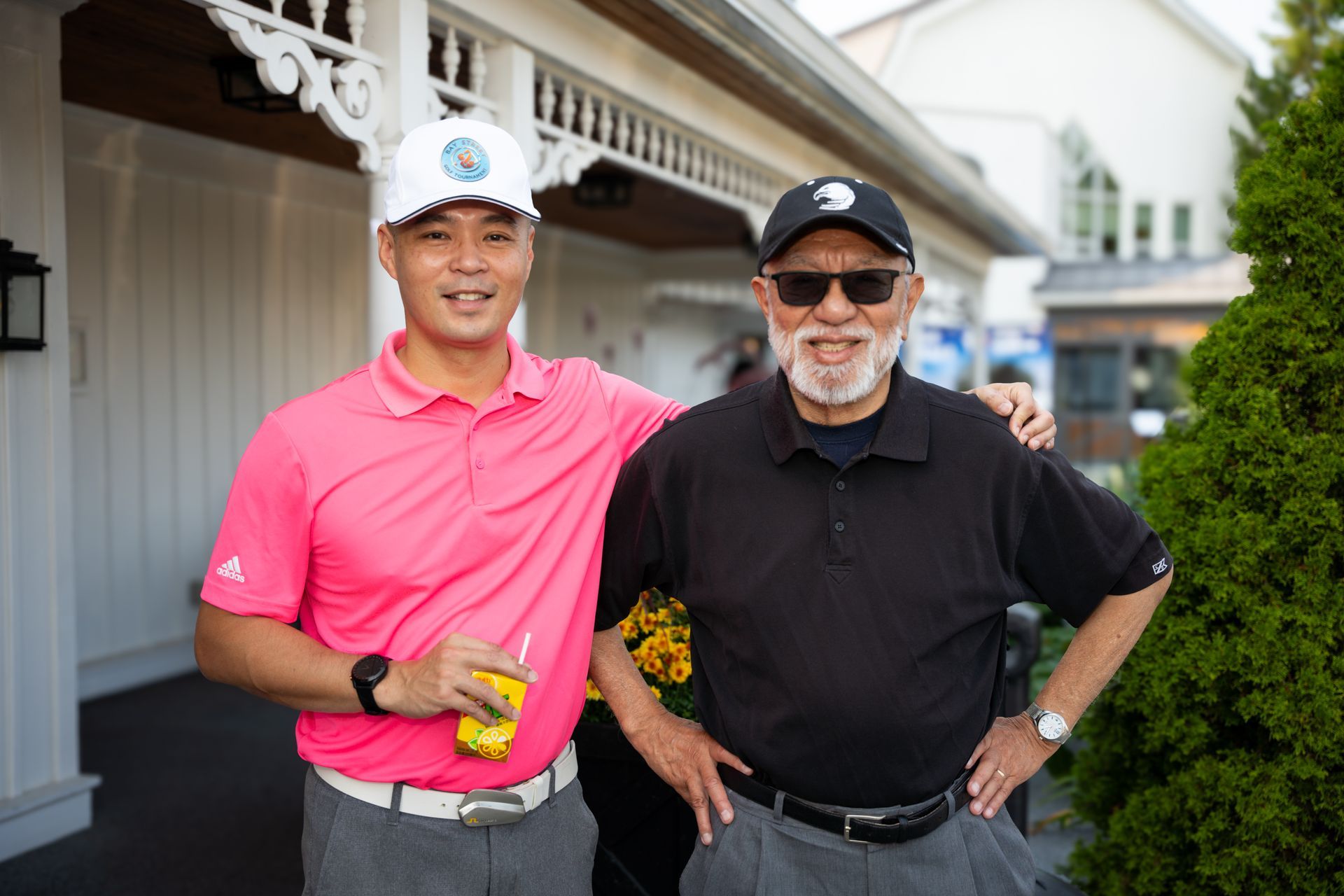 Two men in hats and casual attire pose outdoors, one with arm around the other.