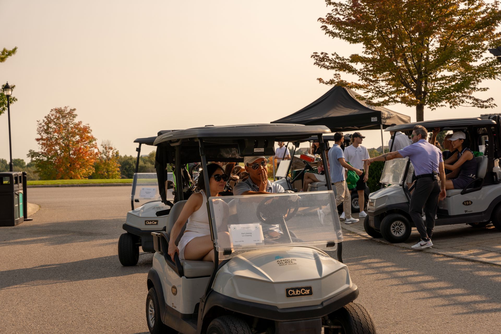 Golf carts with people on a course. People gather near a tent; a person in a purple shirt is directing. Sunny day.