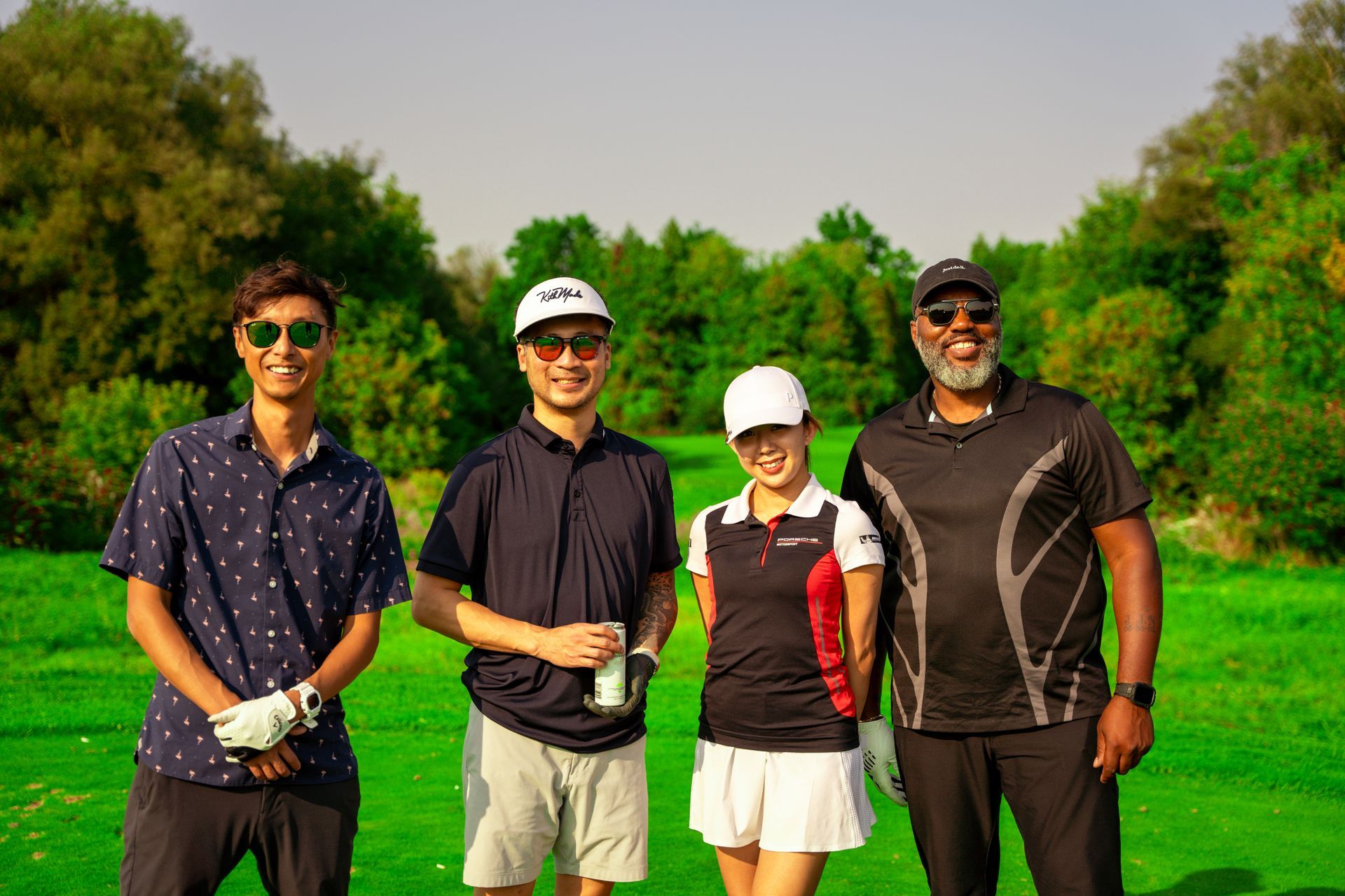Four people posing on a golf course. Green grass, trees. Cloudy sky. Two men, two women wearing golf attire.