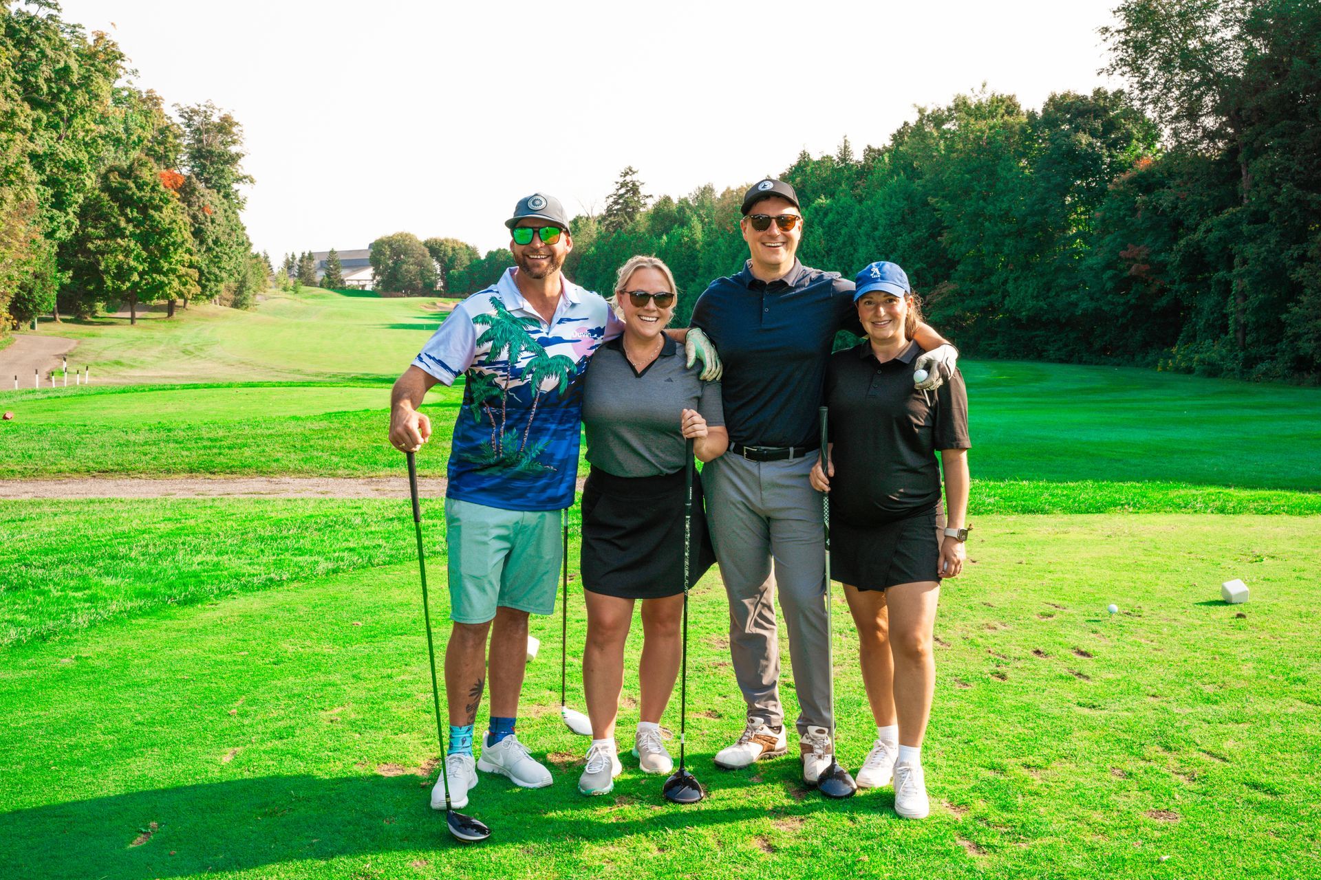 Four people pose on a golf course. Two men and two women stand on a green, smiling. The sky is bright.