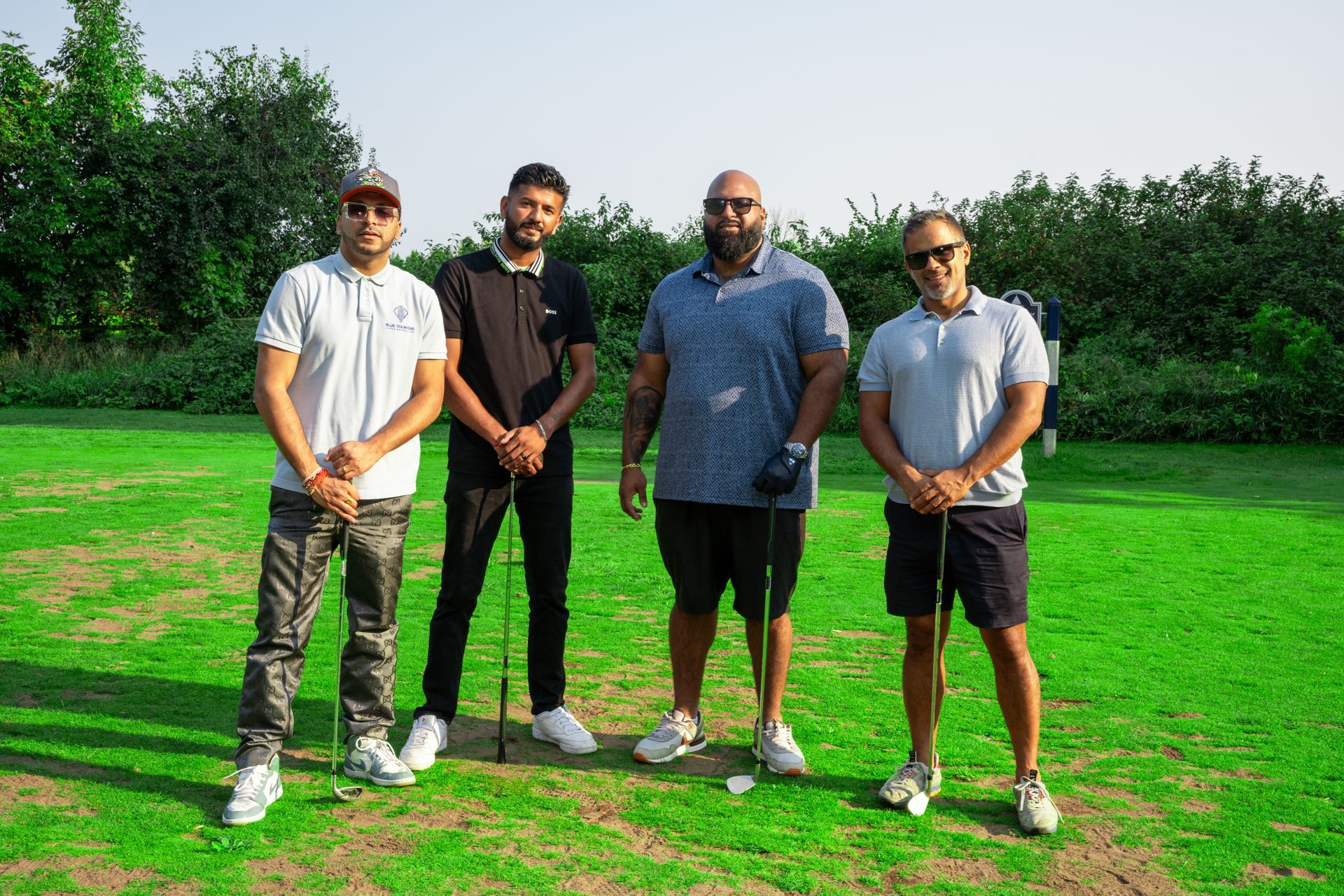 Four men standing on a golf course holding golf clubs. Green grass, trees, and bright daylight.