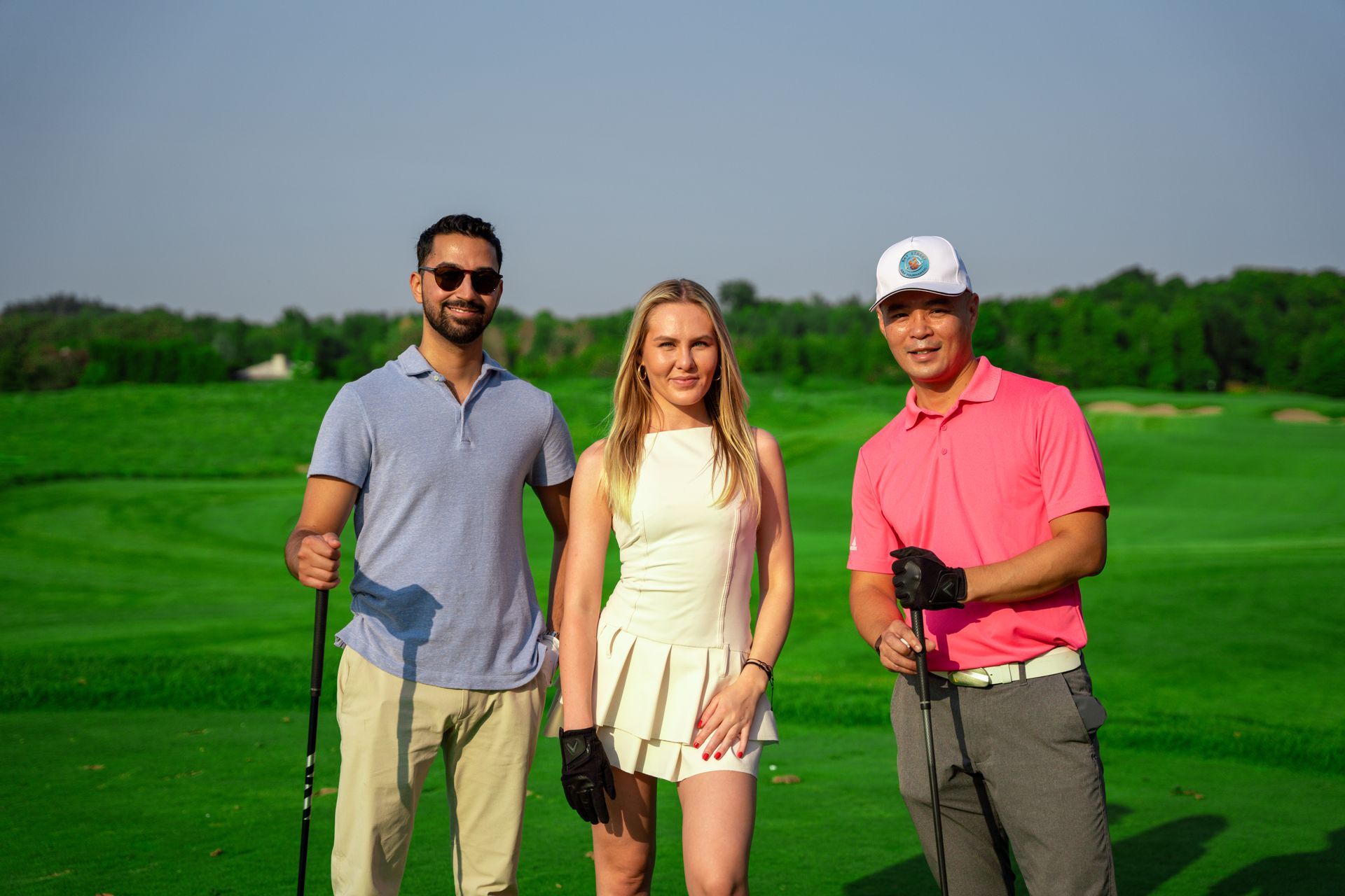 Three people on a golf course, smiling and holding clubs. Green grass, blue sky, sunny day.