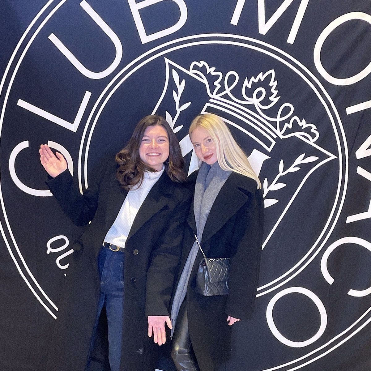 Two people pose in front of a “Club Monaco” logo. One wears a black coat, the other, a coat and scarf.