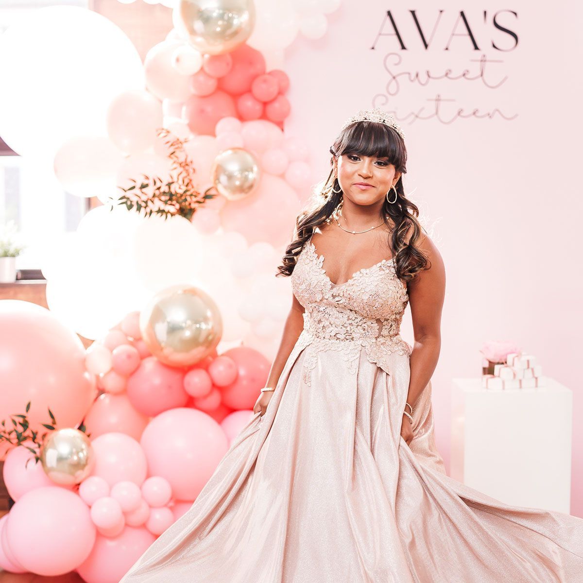 Teenager in formal gown poses in front of a pink balloon display celebrating her Sweet Sixteen.