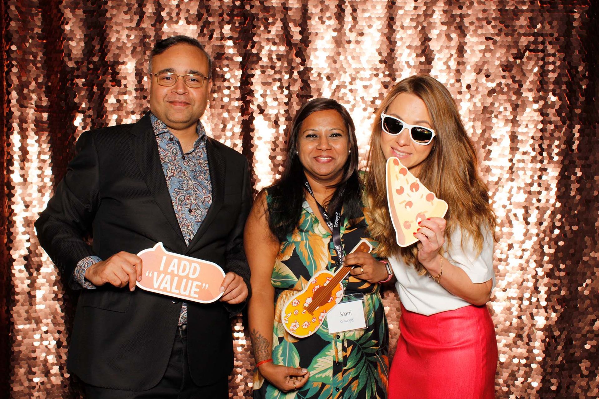 Three people pose for a photo booth with props against a sequined backdrop.