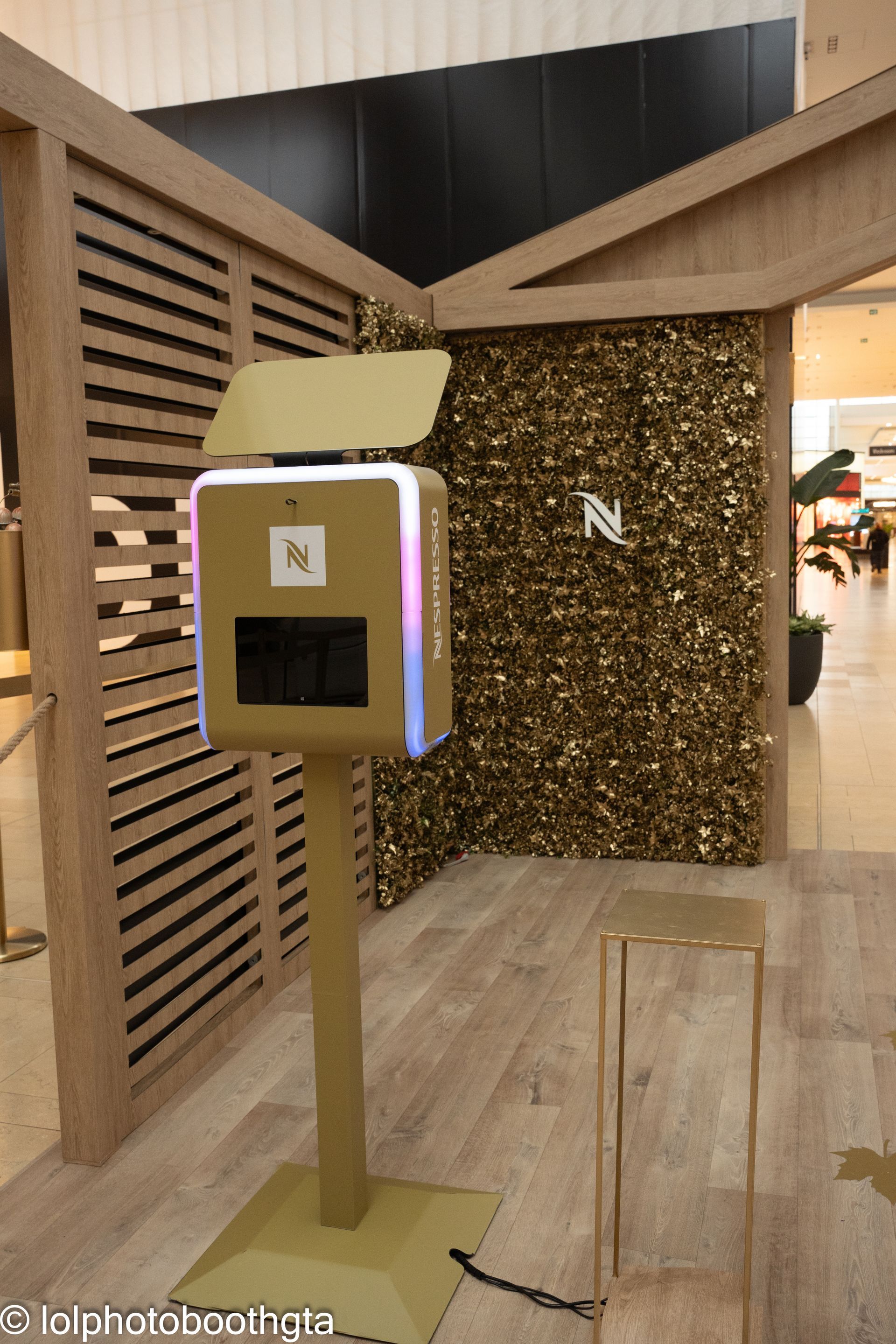 Photo booth in a gold and wooden structure, with a backdrop of gold foliage, in a mall setting.