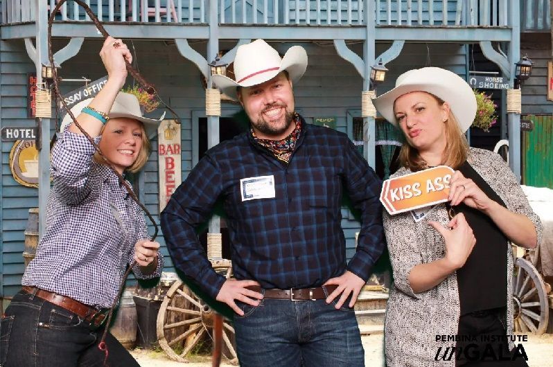 Three people in cowboy attire pose in front of a Western-themed backdrop, one holding a whip, another with a 
