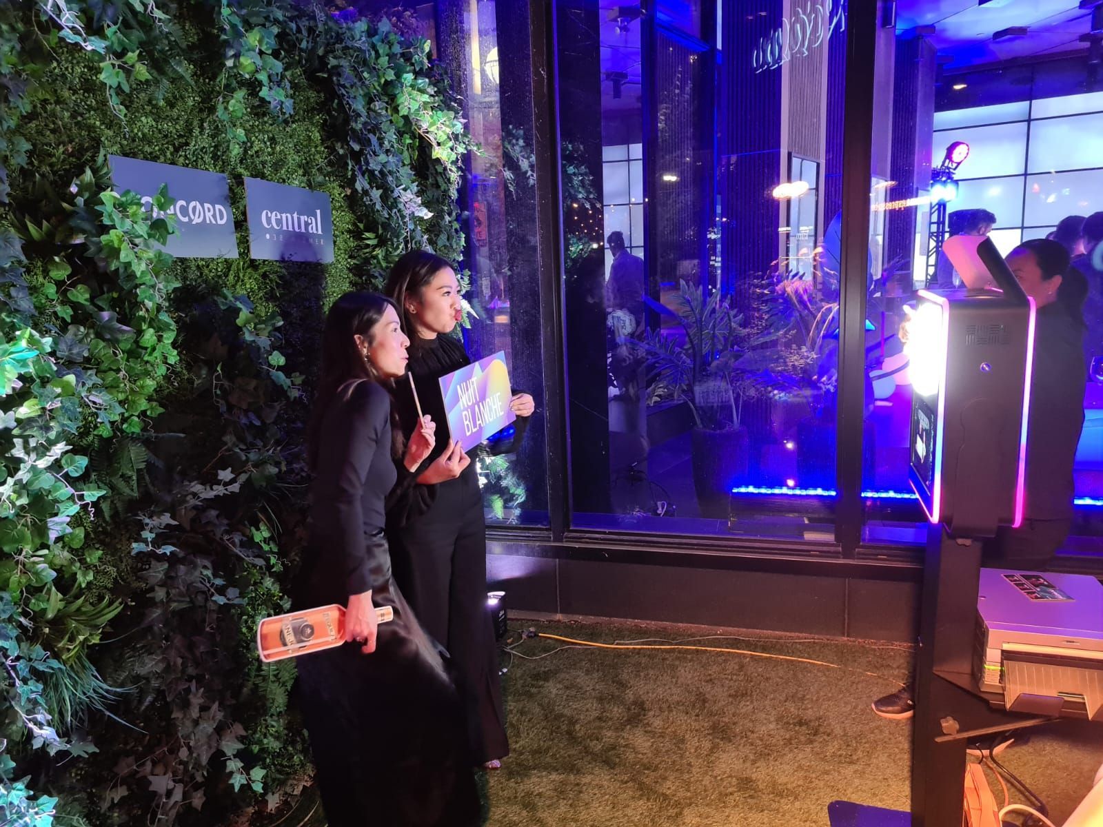 Two people pose near a photo booth with a green wall backdrop. A building is visible in the background.