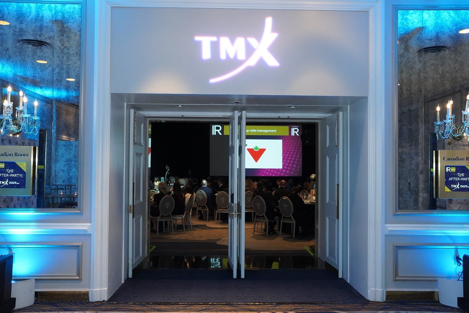 Entrance to a TMX event, featuring a sign and open doors to a gathering with a large screen.