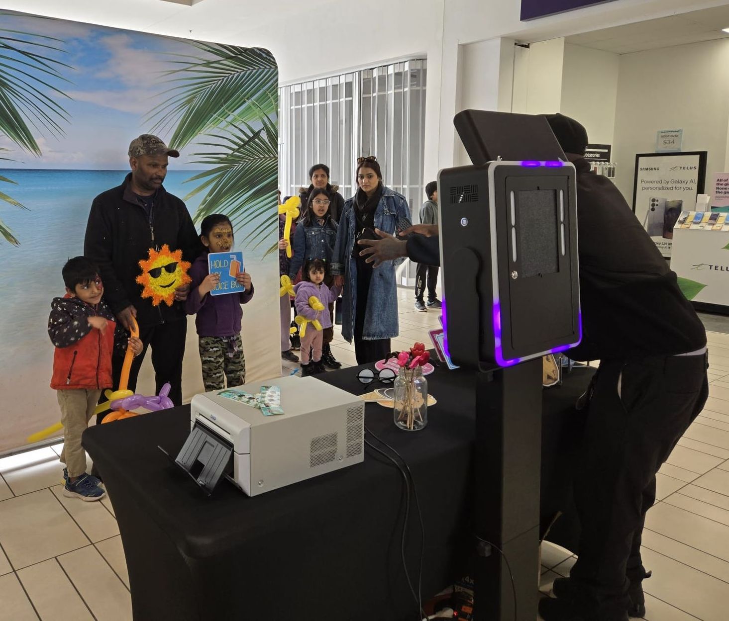 Photo booth with group of people, including children, posing in front of a beach backdrop. Black table, printer, operator.