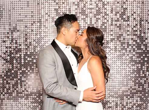 Couple kissing in front of a silver sequined backdrop; man in gray suit, woman in white dress.