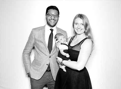 Family of three poses for photo; parents smile, holding a newborn baby. Black and white.