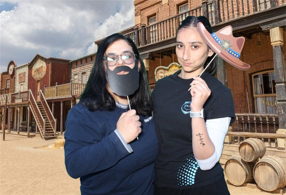 Two people posing with props in front of a Western town backdrop. One holds a beard, the other a cowboy hat.