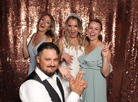 Four people in a photo booth with a copper sequin backdrop. Woman in a wedding dress, two bridesmaids, and a man posing.