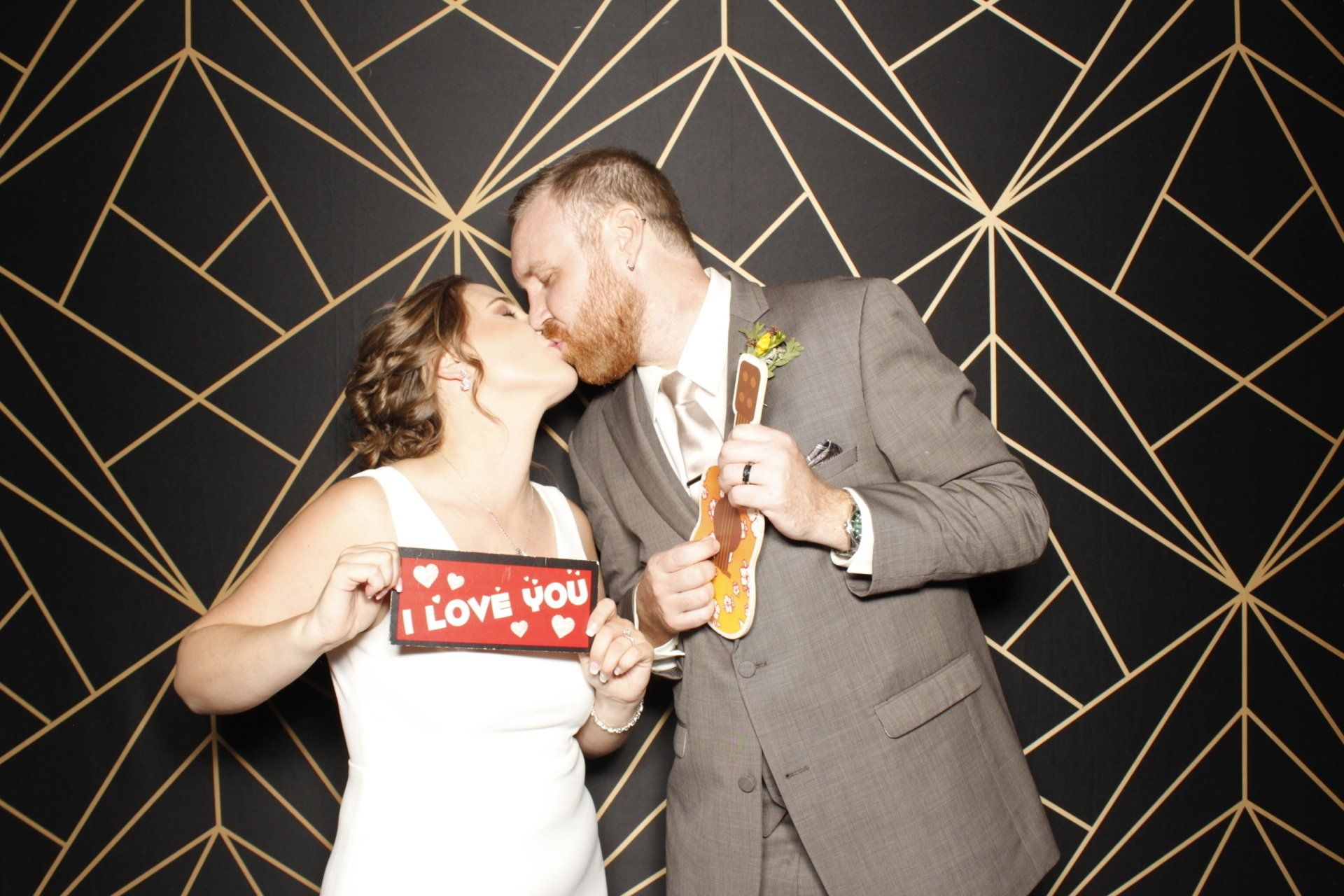 Couple kissing in photo booth, holding props. Bride in white dress, groom in suit, backdrop of gold geometric shapes on black.