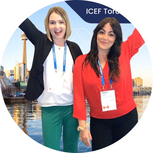 Two women, arms raised, smiling, in front of Toronto skyline backdrop. One wears red, the other green pants.