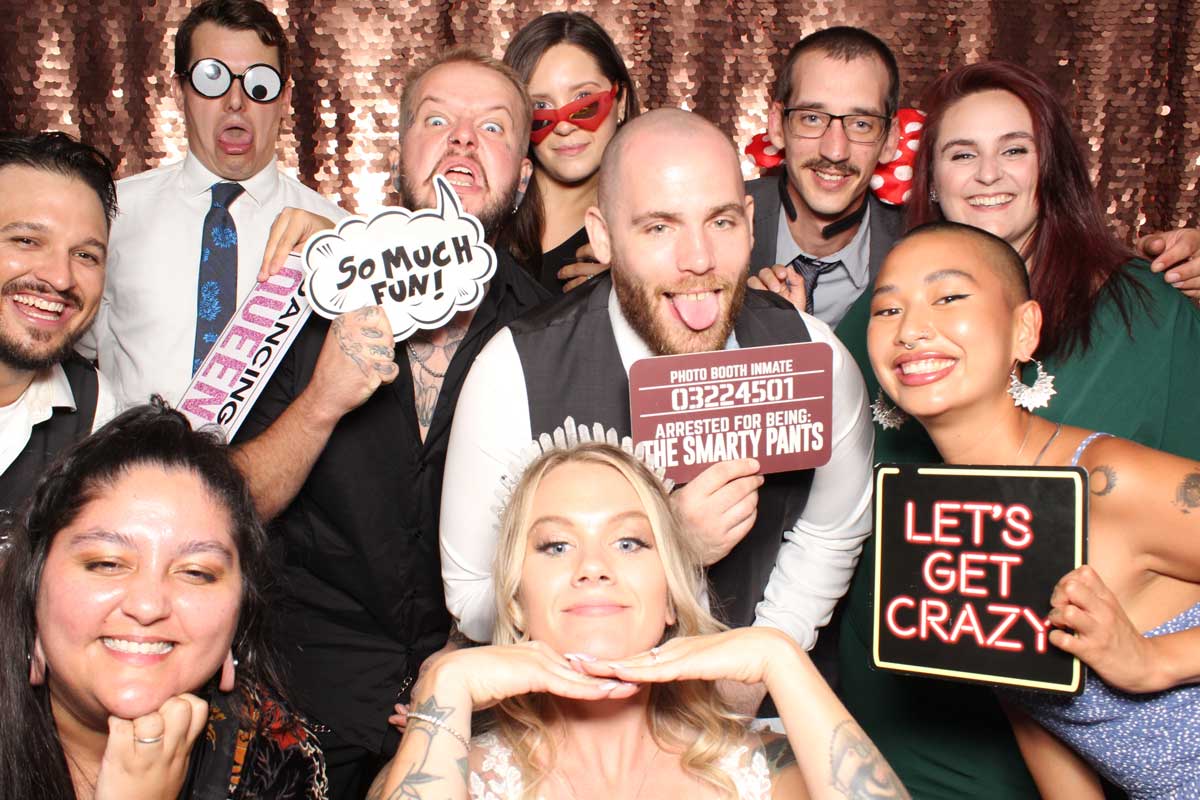 Group photo booth shot with people smiling and posing with props against a rose gold sequin backdrop.
