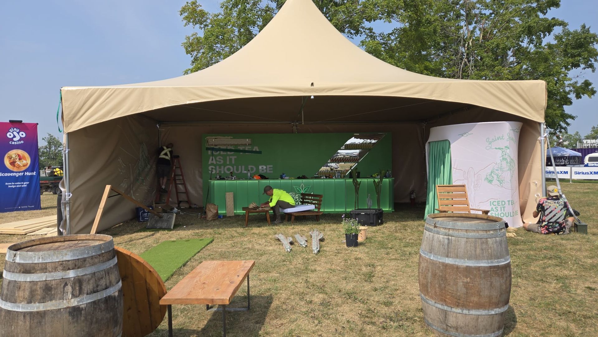 Beige tent set up outdoors for an event. People working on a green backdrop, surrounded by barrels and a bench.