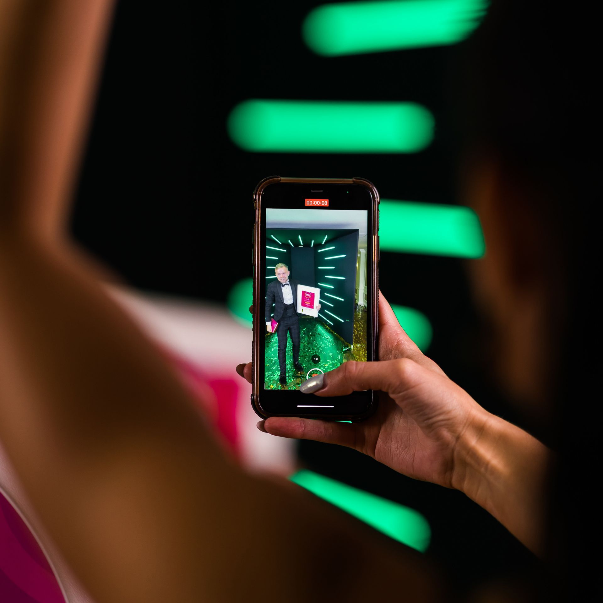 Person recording a man on a phone screen, who is holding a framed award in front of a green-lit background.