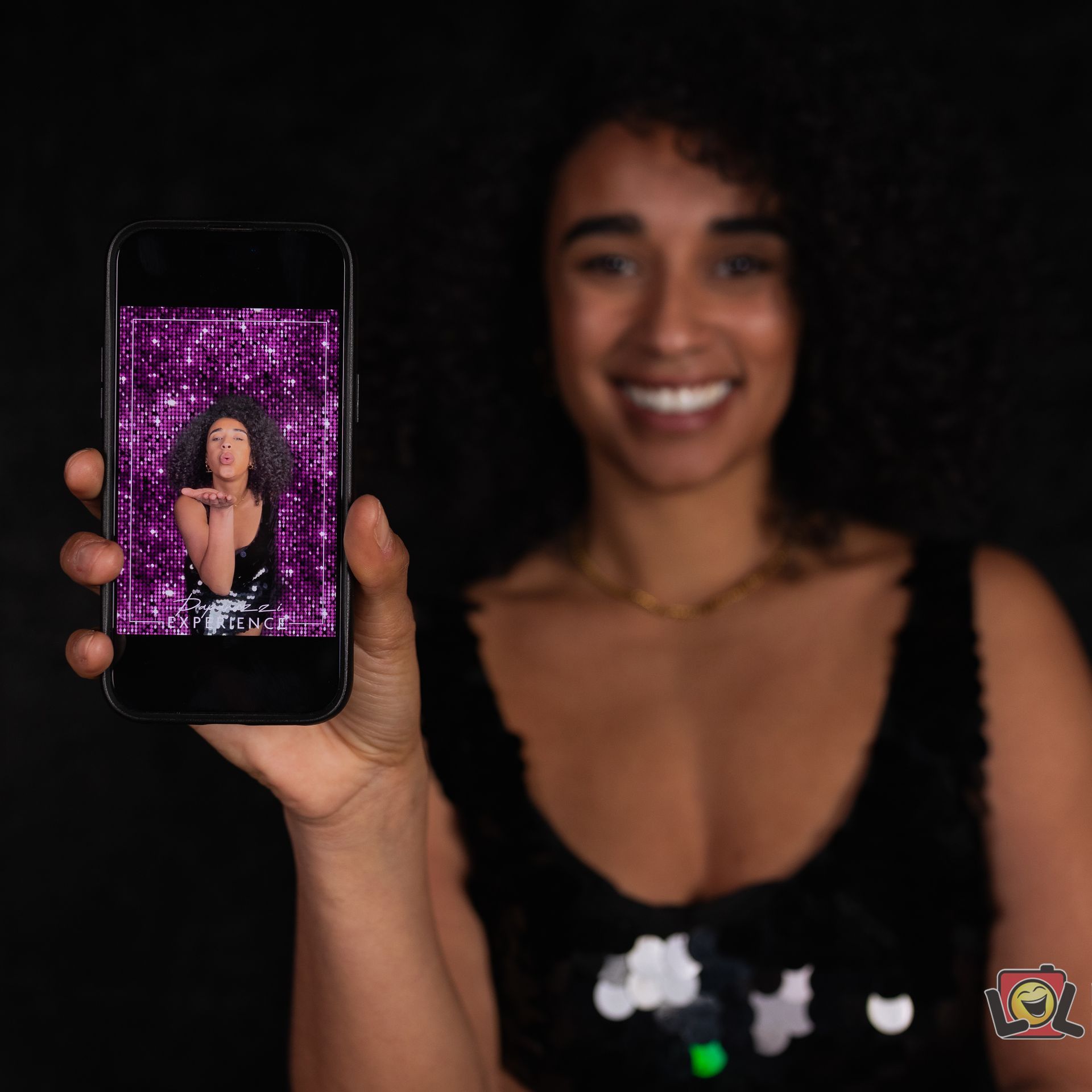 Woman smiling, holding phone showing photo of herself in front of a purple backdrop.
