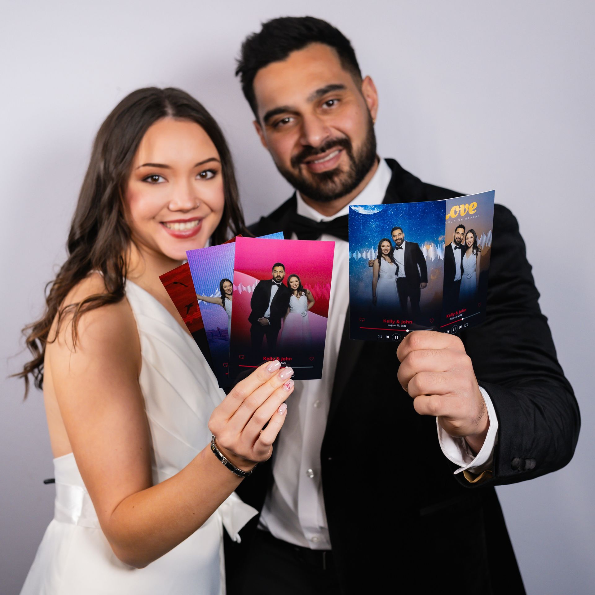 Couple smiling, holding photo booth pictures. Man in tuxedo; woman in white dress. Studio setting.