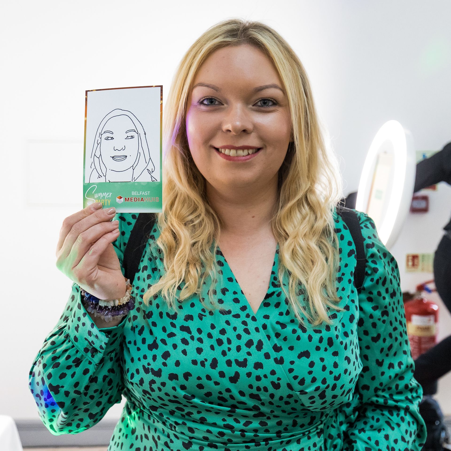 Woman in green dress holds a drawing of a face; smiling; indoors with ring light and fire extinguisher.