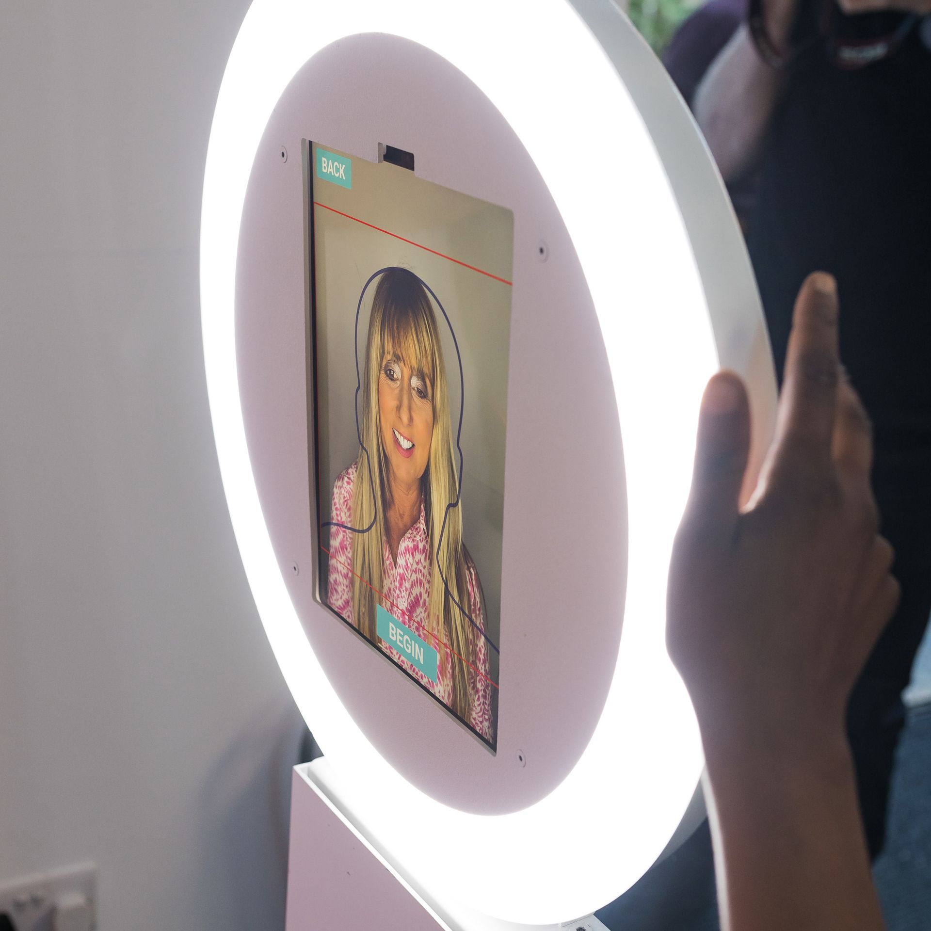 Person interacting with a photo booth, illuminated by a ring light. The booth displays a photo of a person.