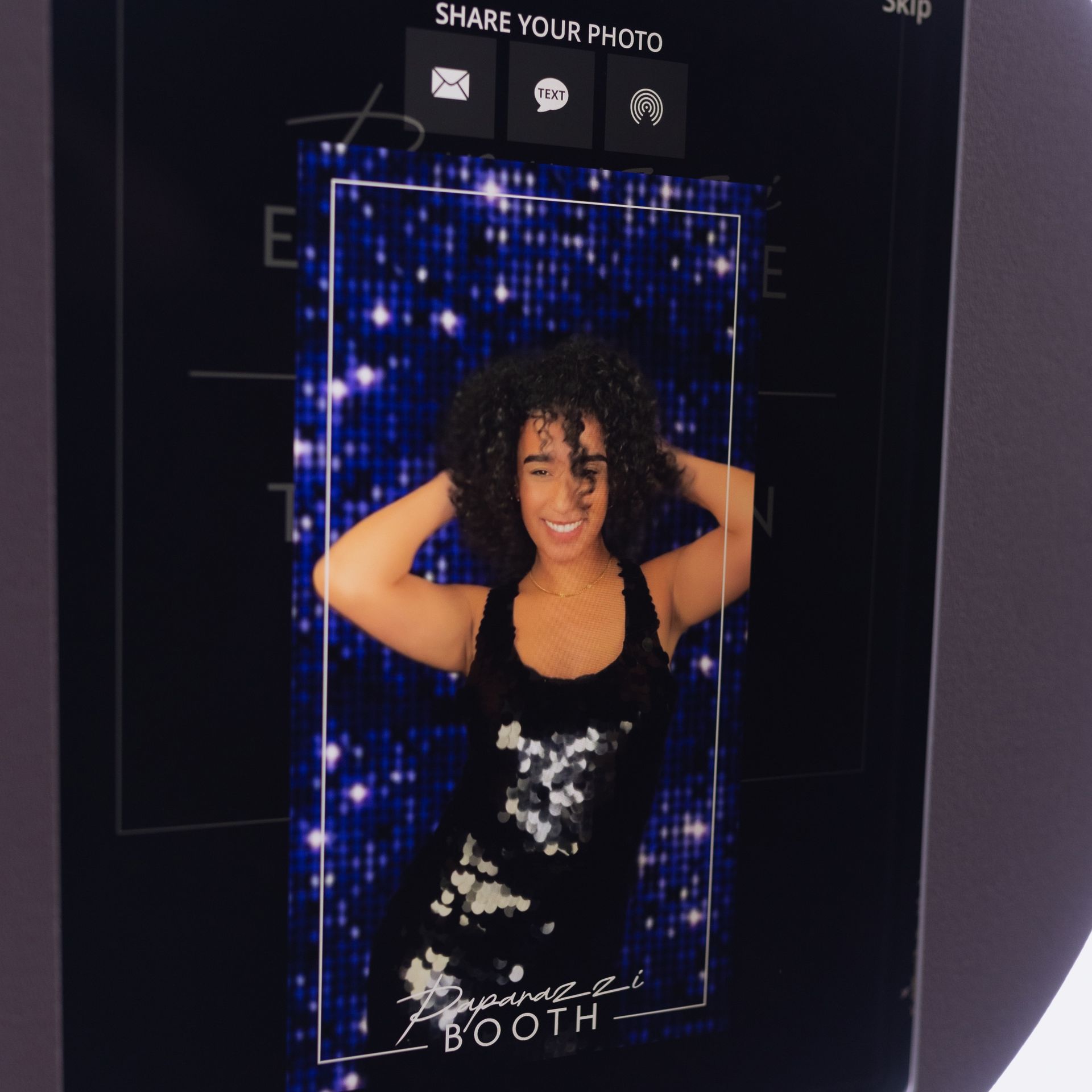 Woman in a photo booth smiling with hands on her head, wearing a black sequined dress. Blue glitter backdrop.