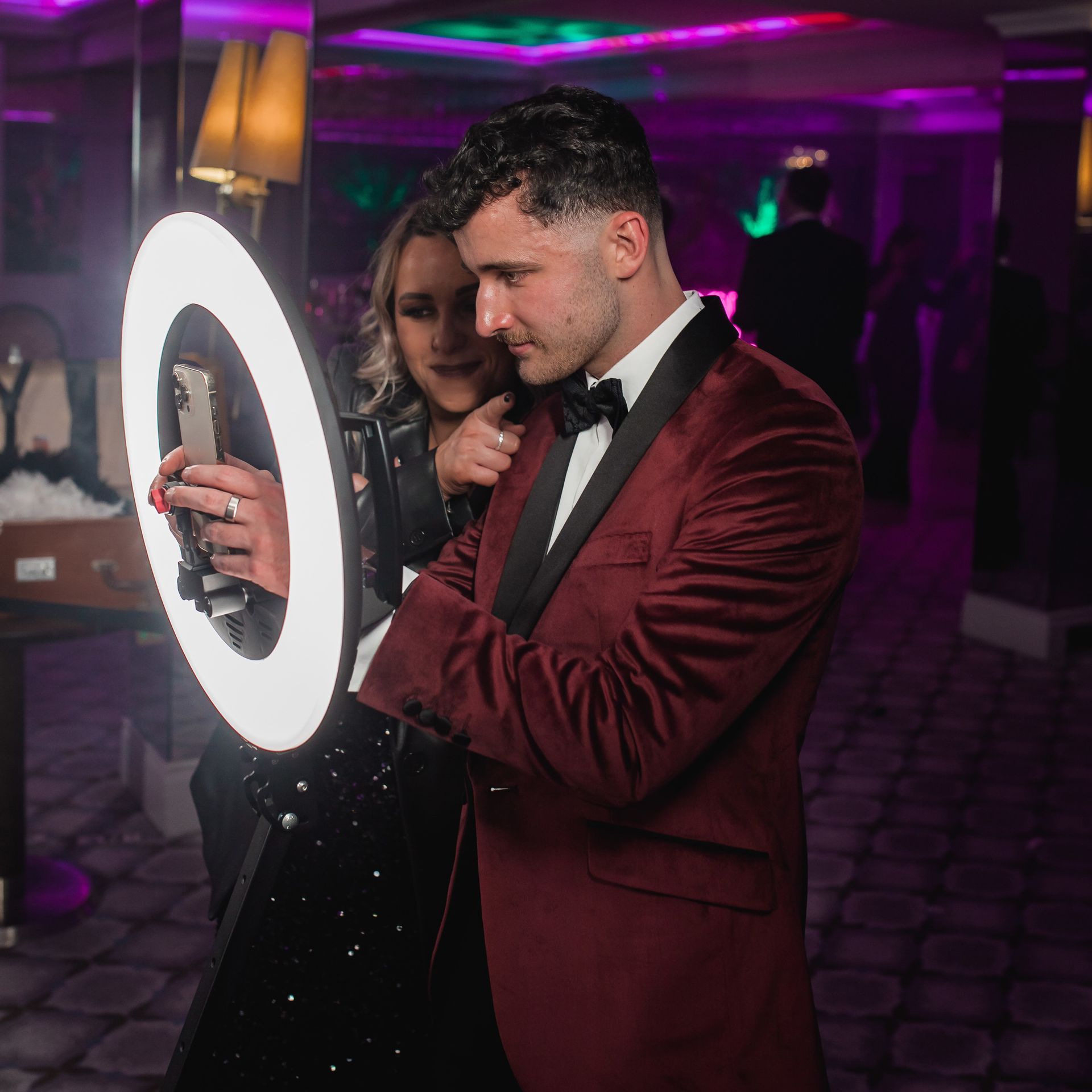 A man in a red suit and a woman pose by a lit photo booth with a ring light in a party setting.