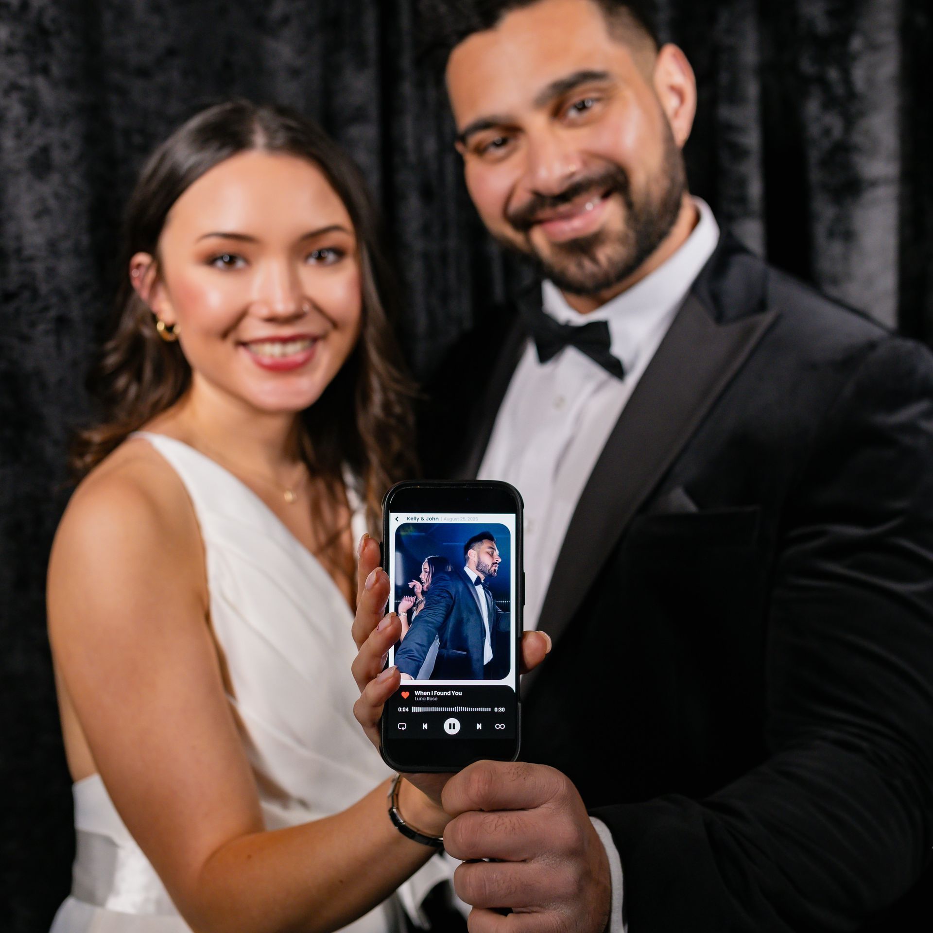 Couple smiles, holding phone displaying photo of a man in tuxedo; formal setting with black backdrop.