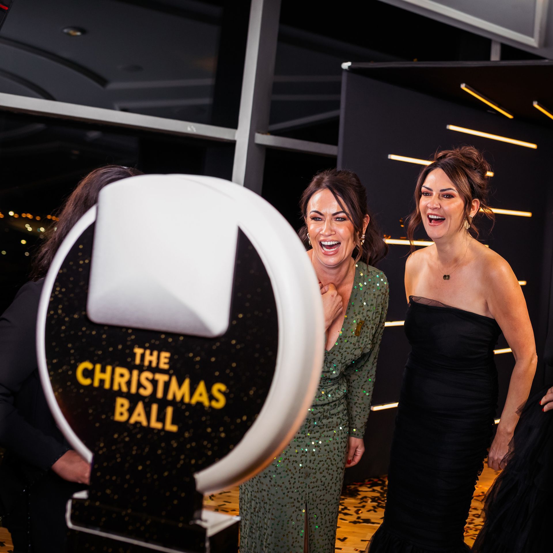 Two women laughing, posing by photo booth at Christmas Ball event. Glittering green and black dresses.
