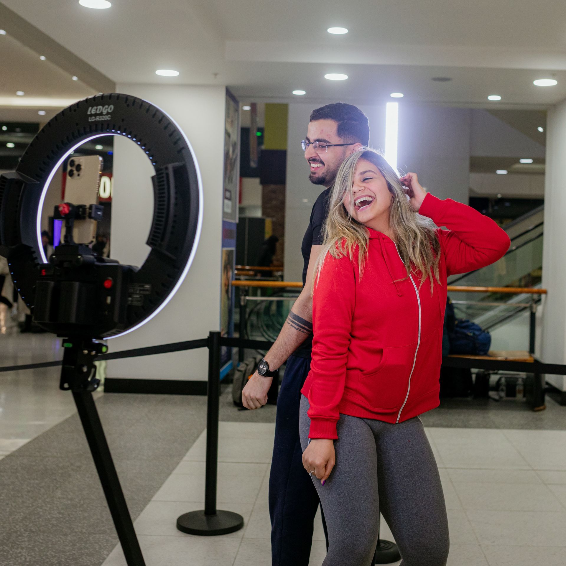 Couple posing for a photo with a ring light in a mall. Woman in red jacket, man in black shirt.