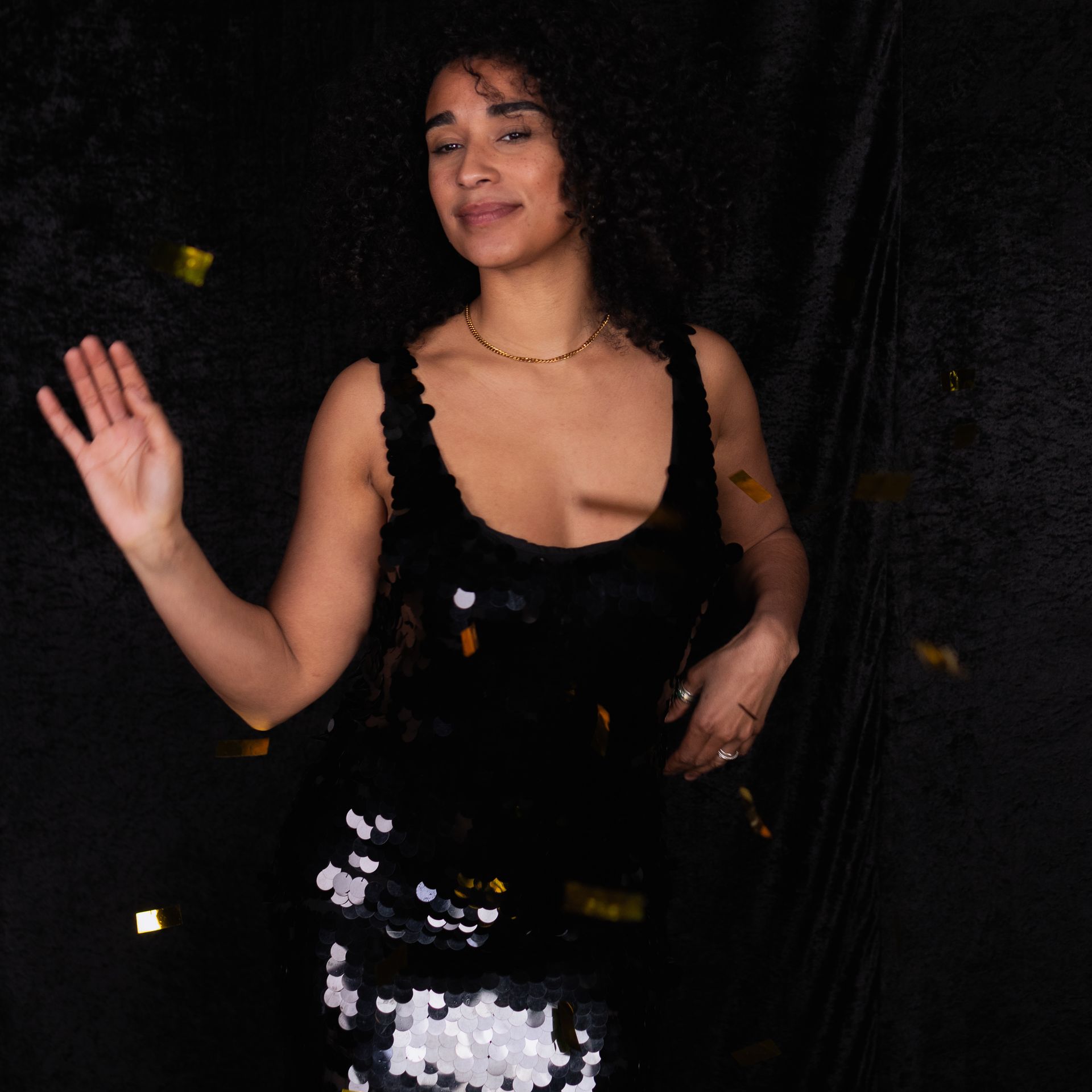 Woman in black sequined dress waves, confetti falling, against dark backdrop.