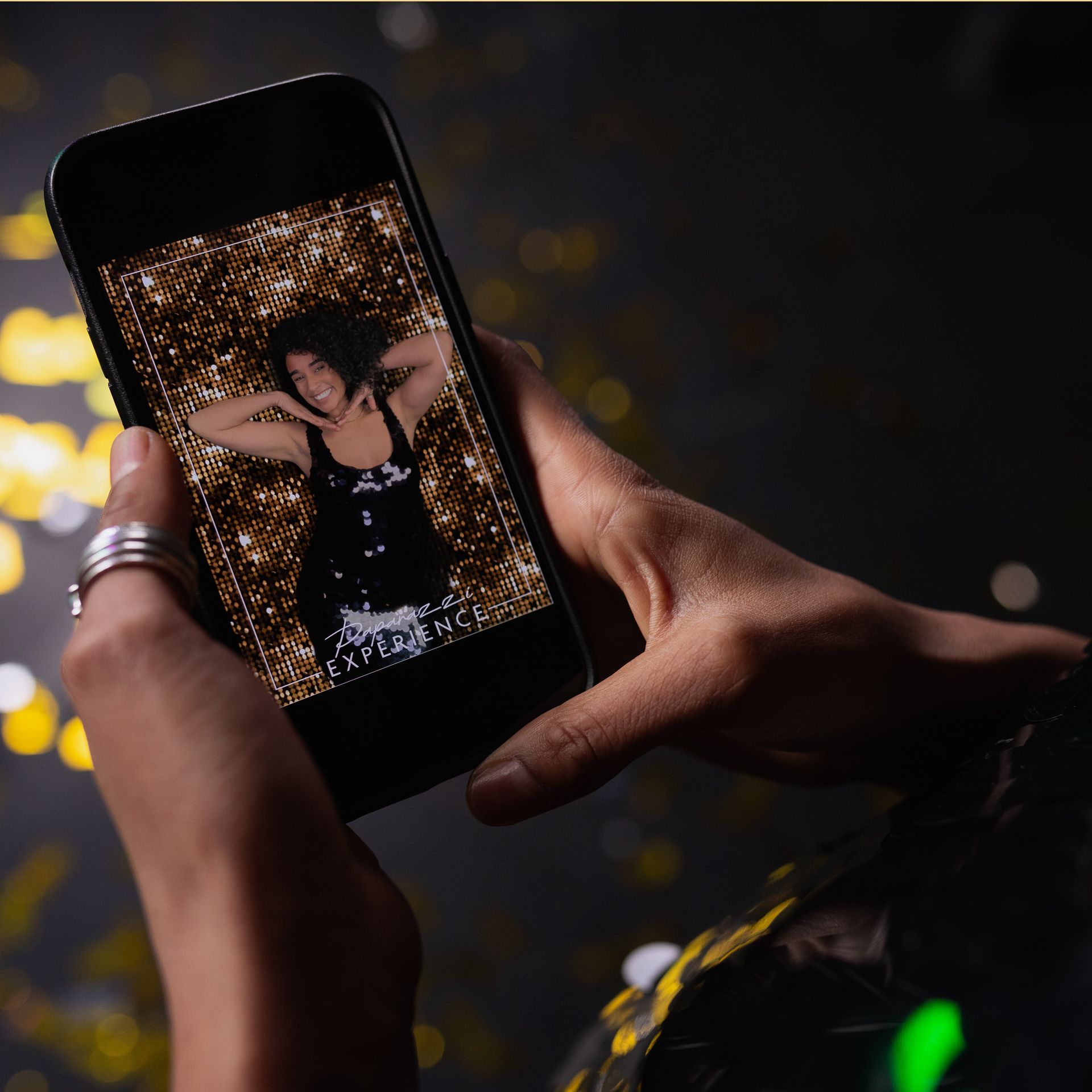Hands holding a phone displaying a photo of a person posing with arms up, against a glittery background.