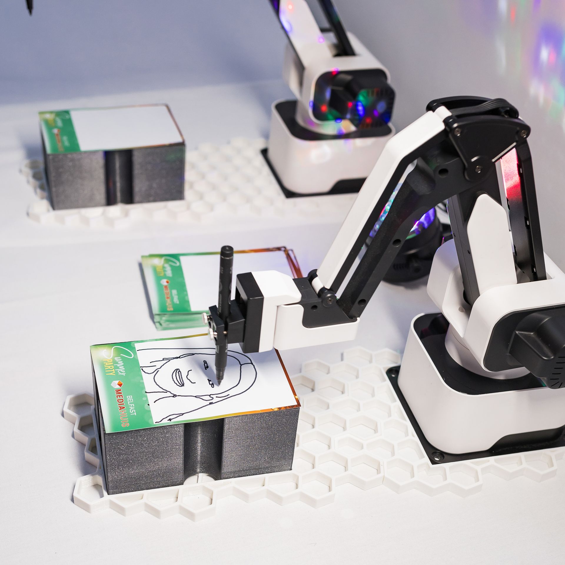 Robotic arm drawing a face on a white card atop a foam block, another card beside it, in a lab setting.