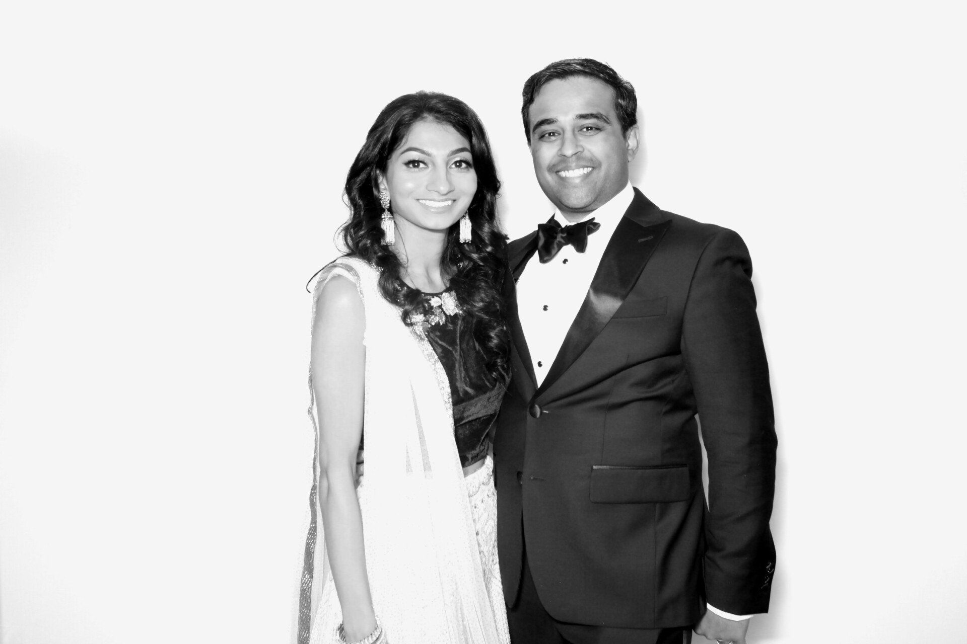 Couple in formal wear smiling against a white backdrop; woman in dress, man in tuxedo.