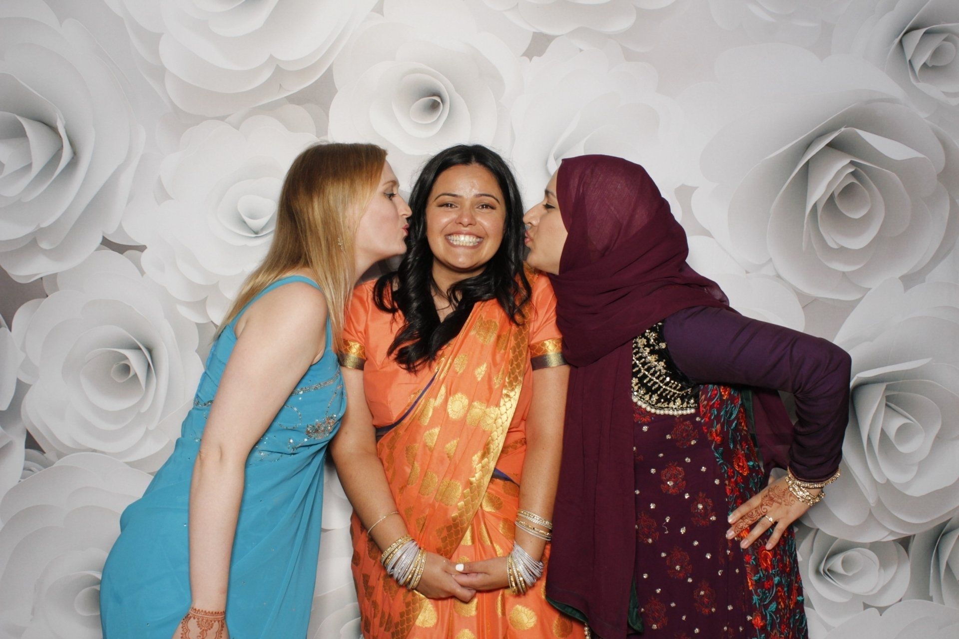 Three women pose for a photo with white rose backdrop; two kiss one woman on her cheeks.