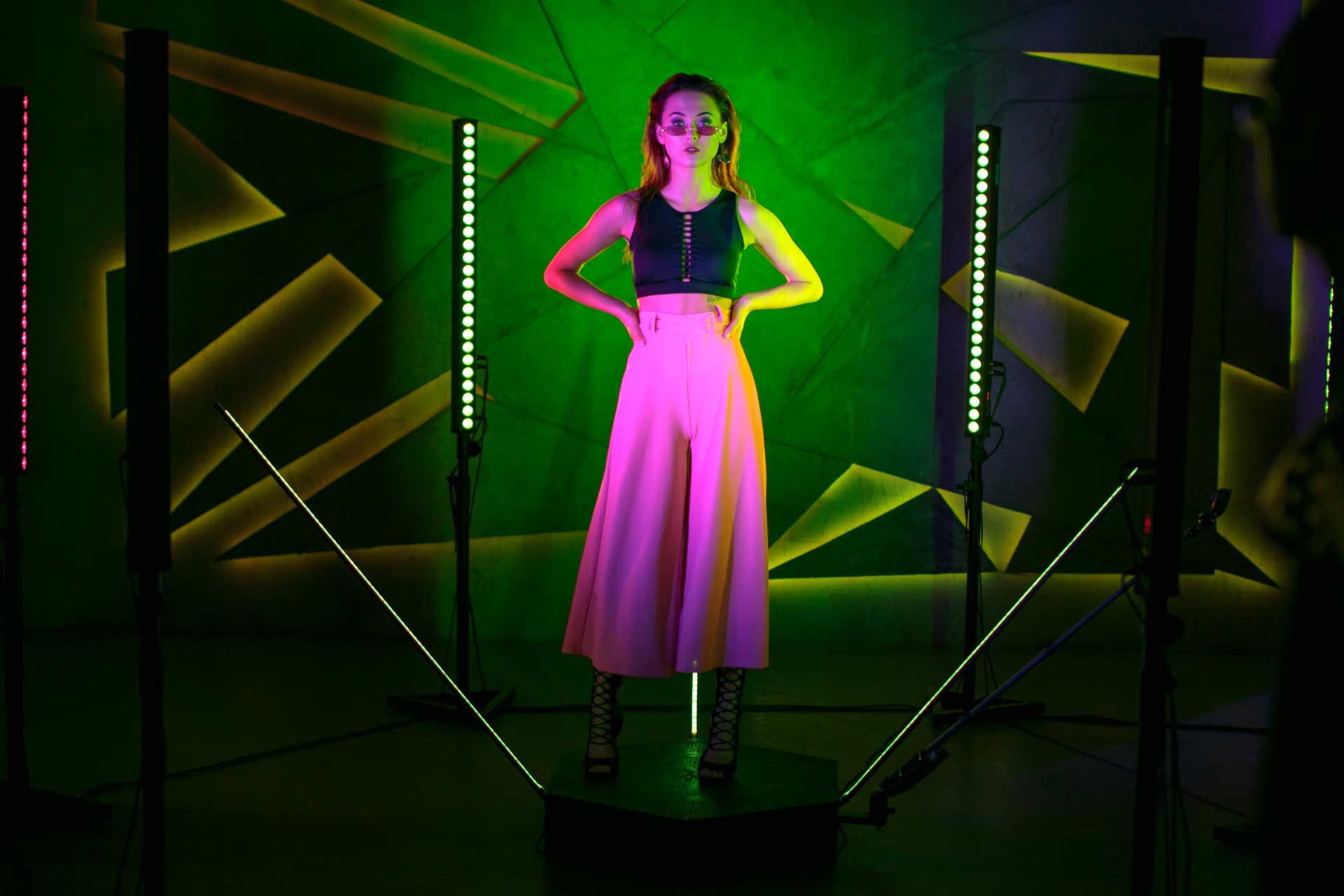 Woman in pink pants and black top poses in a brightly lit studio, arms on hips.