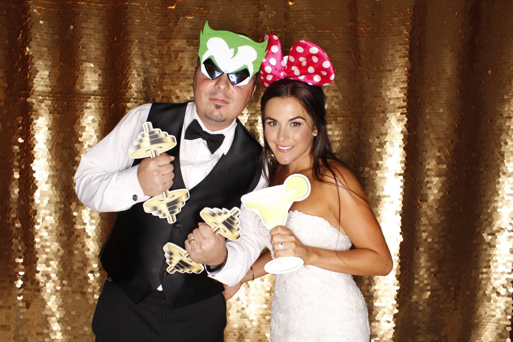 Couple in wedding attire posing with props, gold sequin backdrop. Man in Joker mask, woman with bow, holding drink cutout.