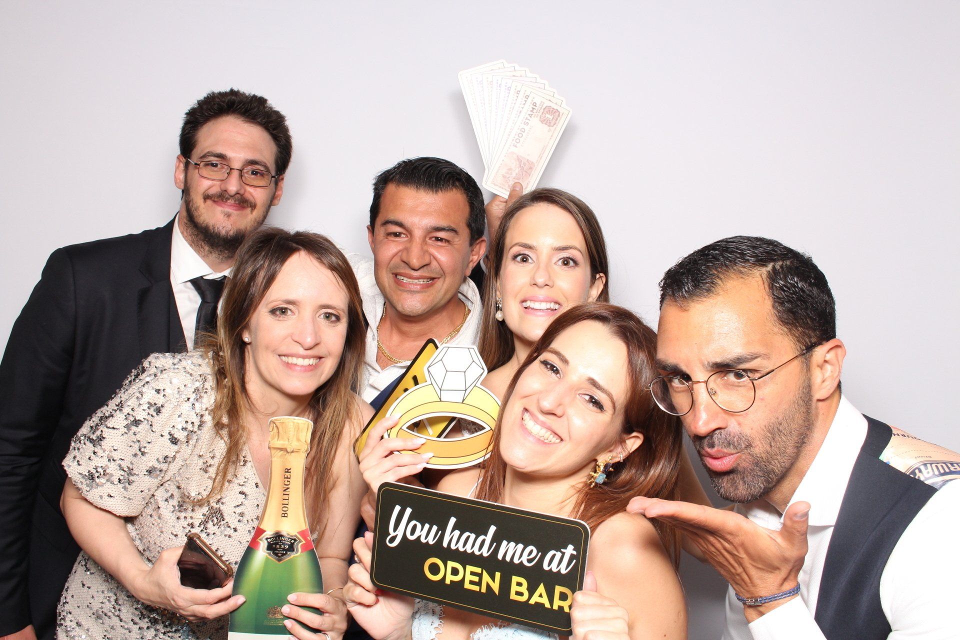 Group of people smiling, posing in a photo booth; holding props like champagne bottle and a sign that says