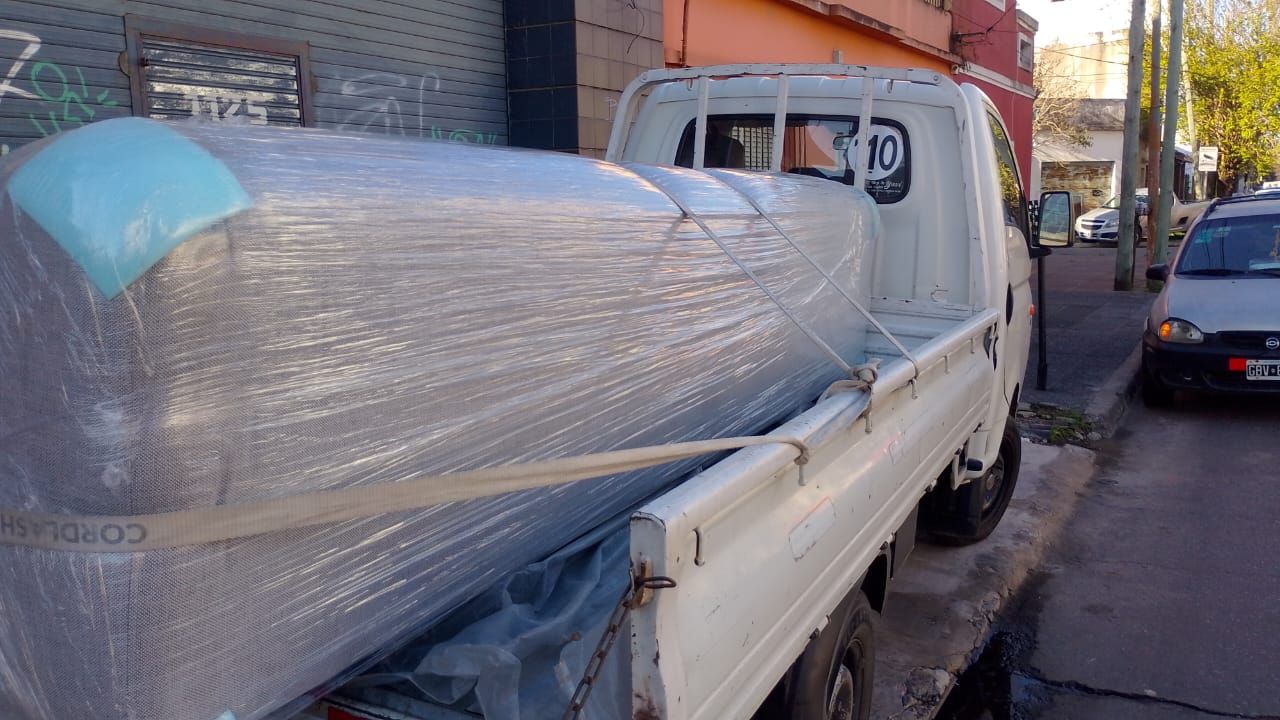 Camioneta blanca transportando un objeto grande envuelto en plástico en una calle de la ciudad con autos estacionados.