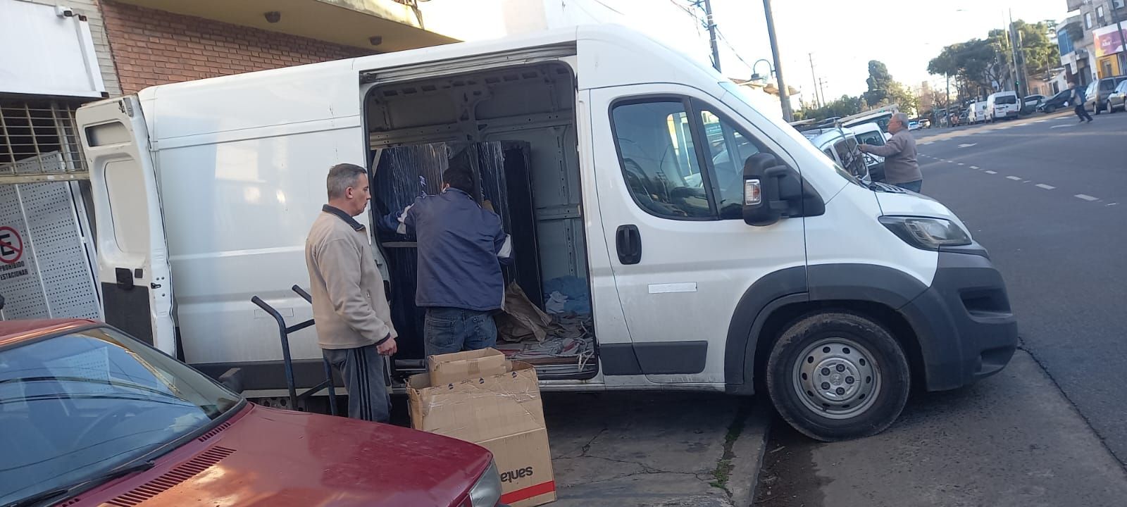 Trabajadores cargando cajas en una furgoneta de reparto blanca en una calle junto a coches aparcados.