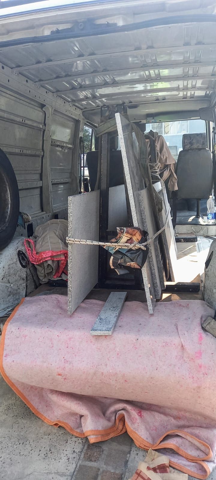 Interior de una furgoneta con tablas apiladas y láminas de metal sujetas sobre un gran cojín rosa.